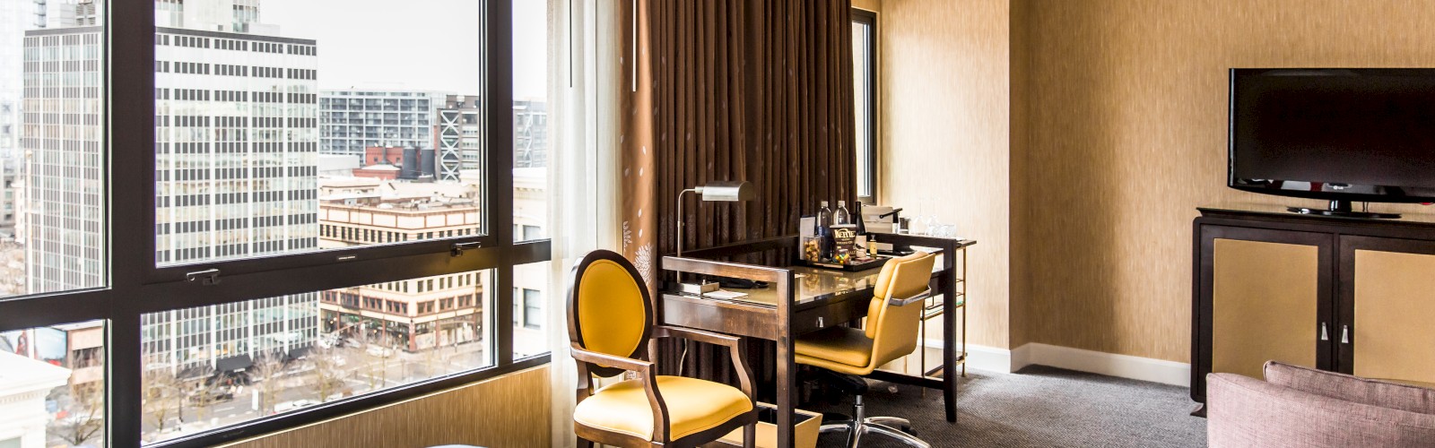 The image shows a hotel room with a large window offering a city view, a bed with blue and red linens, a desk, chairs, and a TV on a cabinet.