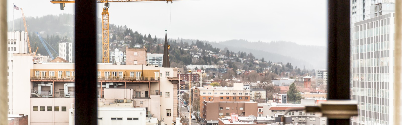 The image shows an urban cityscape viewed through a window, featuring buildings, a construction crane, and a hilly background.