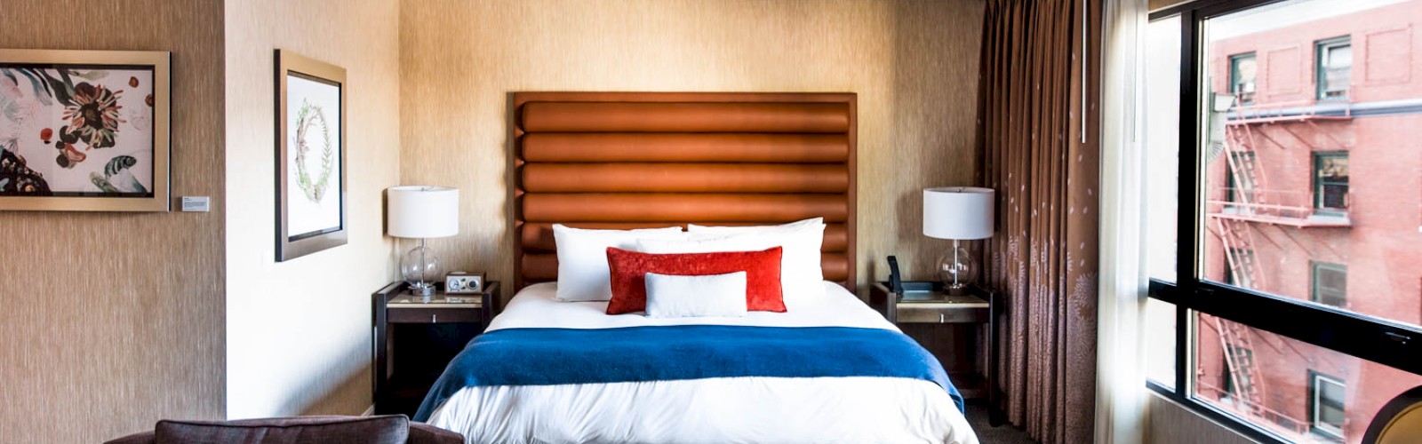 A cozy hotel room features a bed with colorful pillows, a gray armchair, bedside tables, lamps, and large windows with a city view.