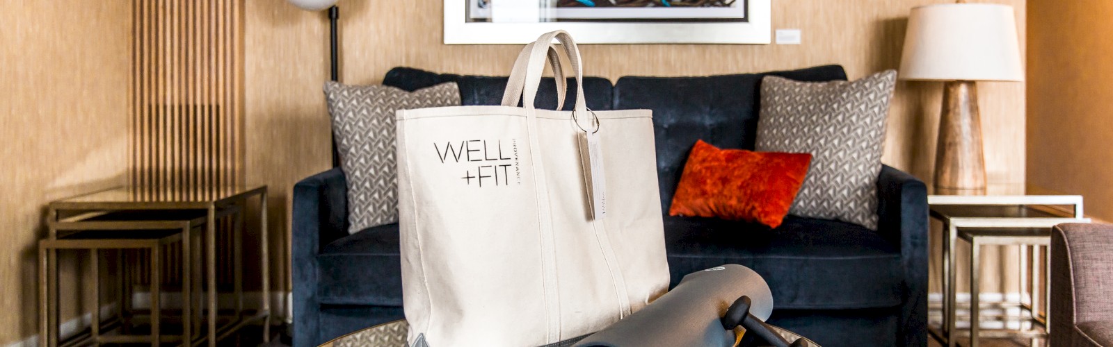 A living room with a black couch, pillows, a wall art, a round coffee table with a Well+Fit bag, yoga mat, and dumbbells.