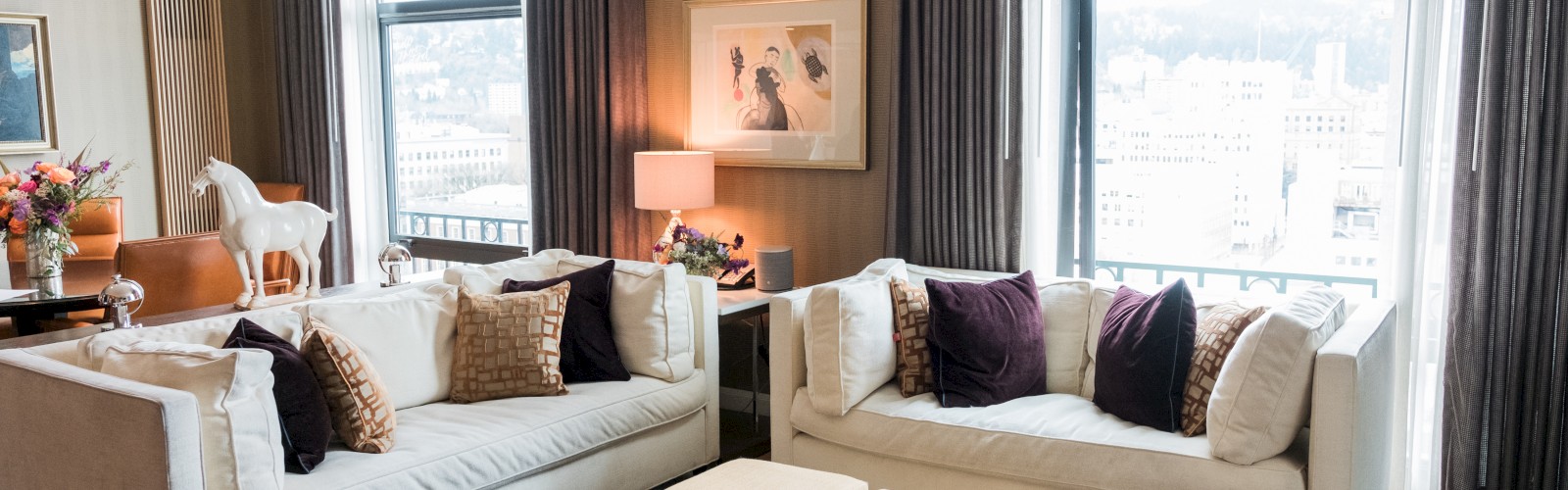 A modern living room with two white sofas, a painting, large windows, purple and brown pillows, and beige ottomans in the center ending the sentence.