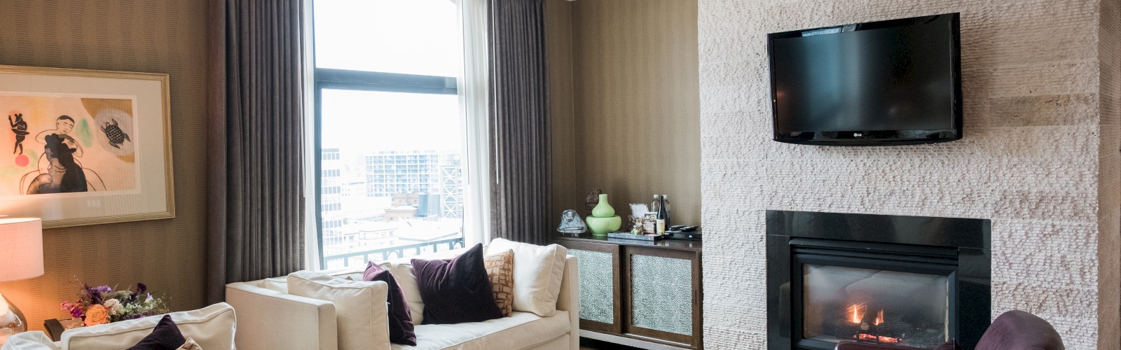 A cozy living room with a fireplace, TV above it, white sofas, brown chairs, a city view window, and wall art.