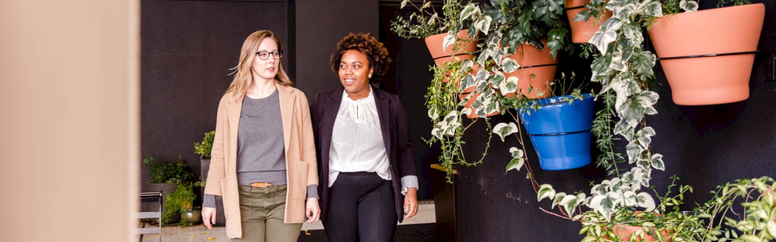 Two women walking beside a vertical garden decorated with various plants in colorful pots, one wearing casual, the other business attire.