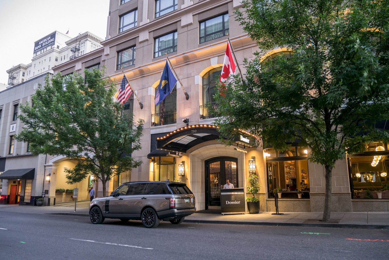 A city street with a luxury SUV parked in front of a building displaying three flags: American, Swedish, and Canadian in the evening.