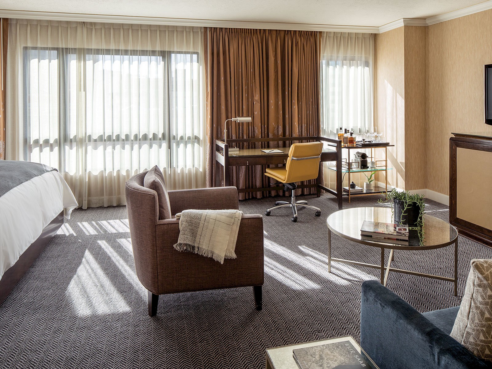 A modern hotel room with a cozy bed, armchair, desk with a yellow chair, minibar, and a coffee table, all lit by large windows with curtains.