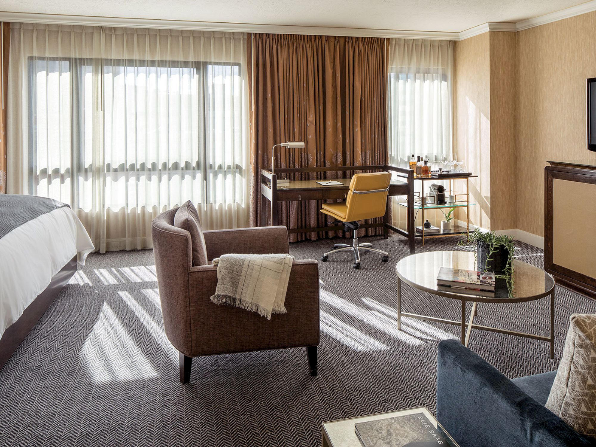 A modern hotel room with a cozy bed, armchair, desk with a yellow chair, minibar, and a coffee table, all lit by large windows with curtains.