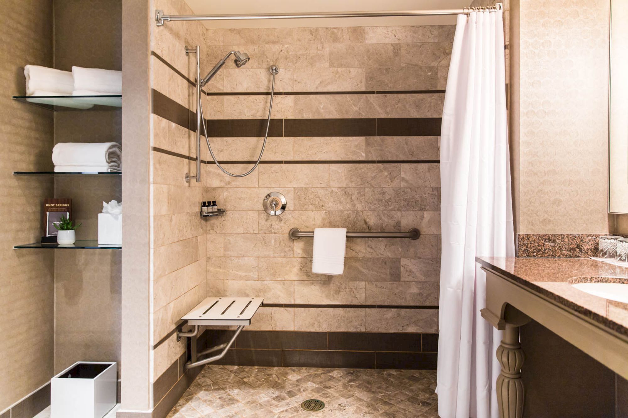 An accessible shower with a fold-out seat, grab bars, and a handheld showerhead. Nearby are shelves with towels and toiletries, ending the sentence.