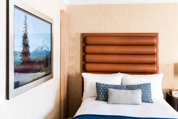 A neatly made bed with blue and gray pillows is next to a wall-mounted landscape painting of trees and mountains in a well-lit room.