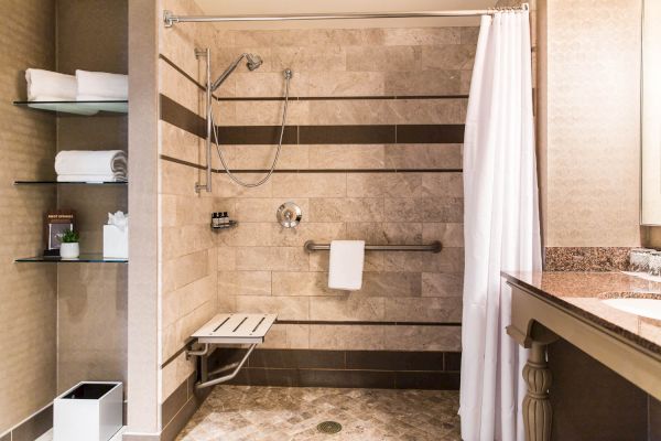 A bathroom features a walk-in shower with a bench, grab bars, and a showerhead. Towels are neatly stacked on shelves on the left wall.