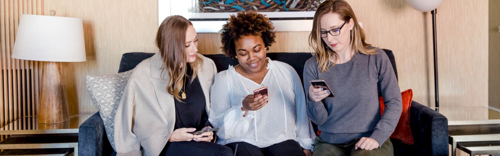 Three people sitting on a couch, looking at their phones. A painting is on the wall behind them. There's a lamp and a side table.