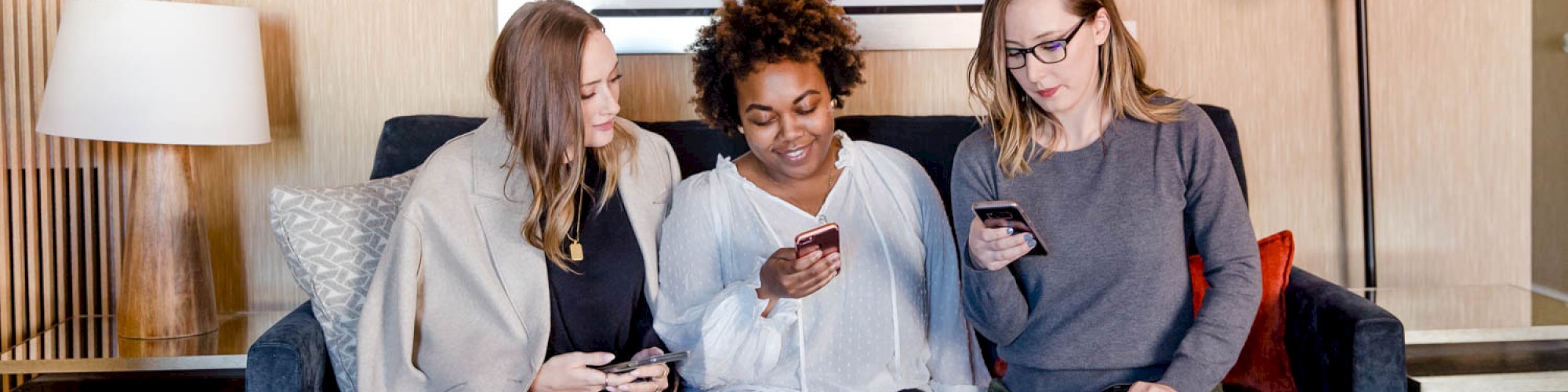 Three people sitting on a couch, looking at their phones. A painting is on the wall behind them. There's a lamp and a side table.