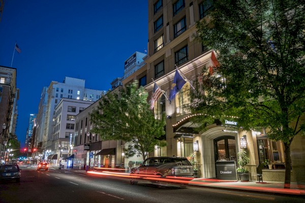 Night city street scene with illuminated buildings, flags, parked cars, and light trails from moving vehicles.