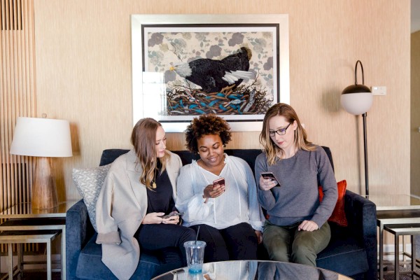 Three people sitting on a couch, looking at their phones, with a framed artwork behind them and a table in front.