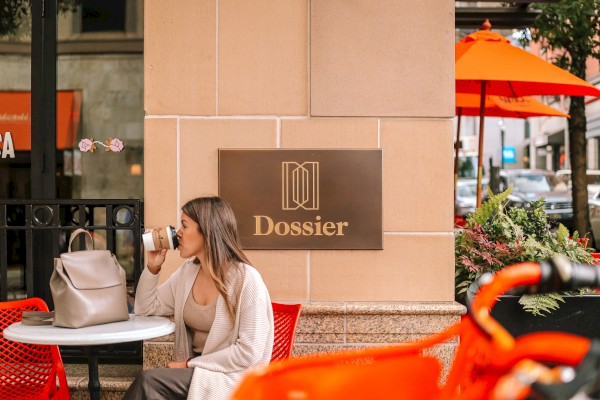 A person sits at an outdoor café table with a backpack, sipping a drink. There's a sign reading 