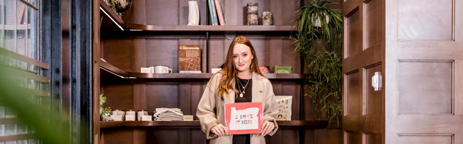 A woman stands in front of a wooden bookshelf holding a framed picture with text, wearing a beige coat, black pants, and patterned boots.