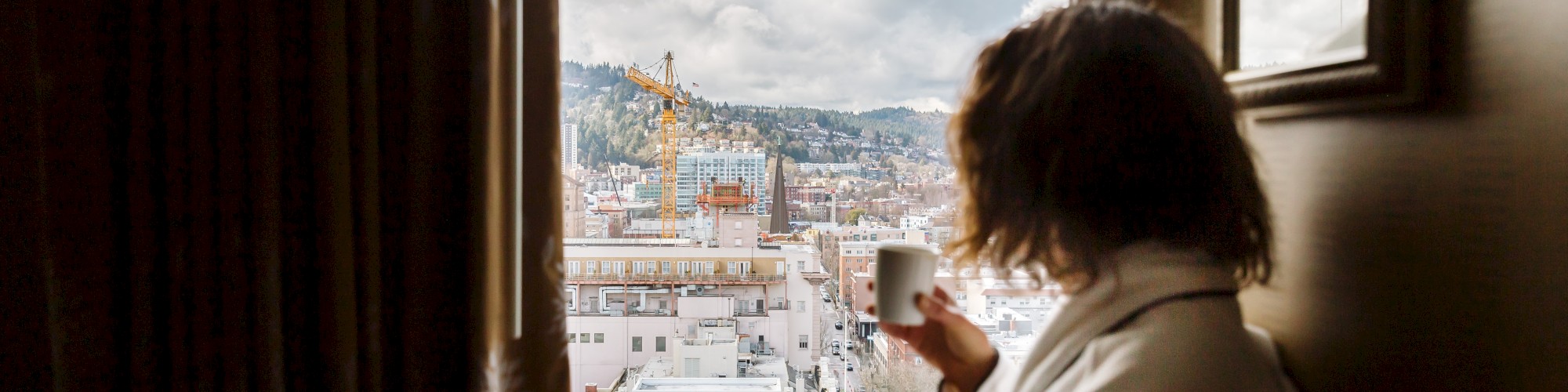 A person in a bathrobe sits by a window, holding a mug, gazing at a cityscape with cranes and buildings under a cloudy sky.