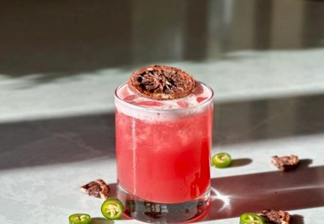 A red cocktail with a garnish, surrounded by chili slices and chocolate pieces.