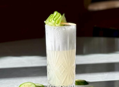 A frothy drink in a crystal glass, garnished with cucumber slices.