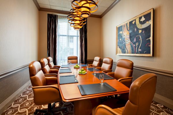 A modern conference room with a long table, brown leather chairs, overhead lights, and a colorful abstract painting on the wall.