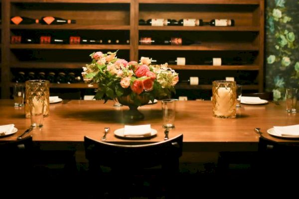 A dining table is set with plates, glasses, and napkins, featuring a floral centerpiece. Behind it are shelves filled with wine bottles.