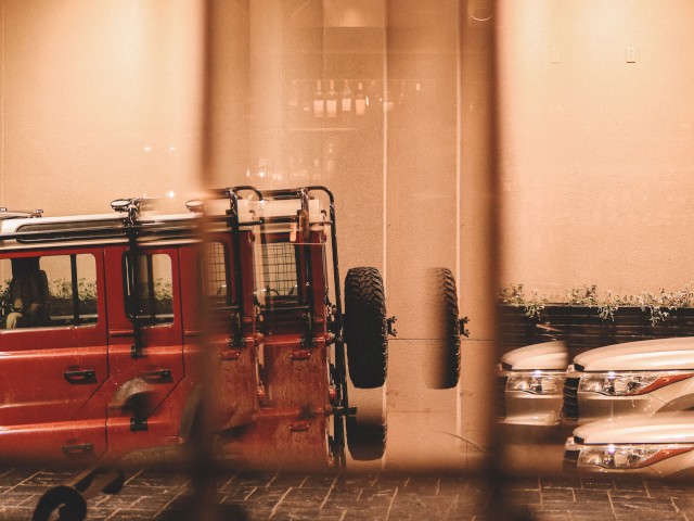 A slightly blurred view through a window showing a red vehicle and two white cars parked in an indoor area with tiled flooring and beige walls.