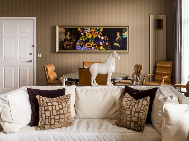 A modern living room with a white couch, patterned pillows, a horse statue, brown chairs, a table, and a painting on the wall with flowers and people.