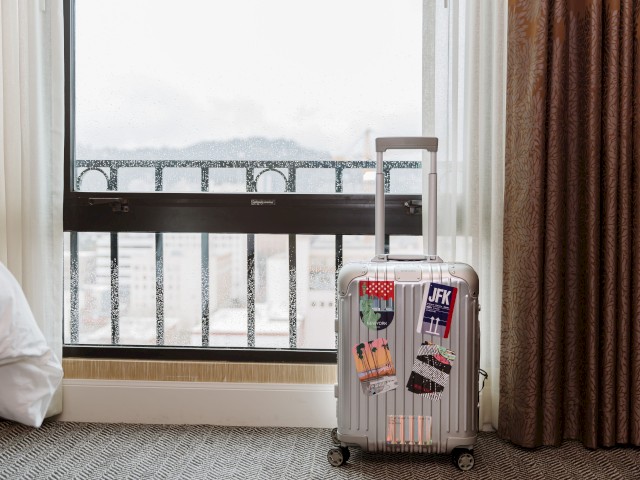 A silver suitcase with travel stickers stands in front of a window, next to a bed and brown curtains in a room with a carpeted floor.