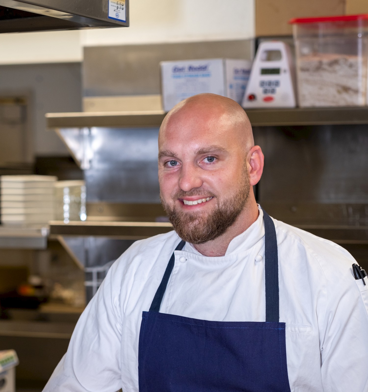 A smiling chef wearing a white coat and blue apron stands in a commercial kitchen, with shelves, containers, and kitchen equipment in the background.