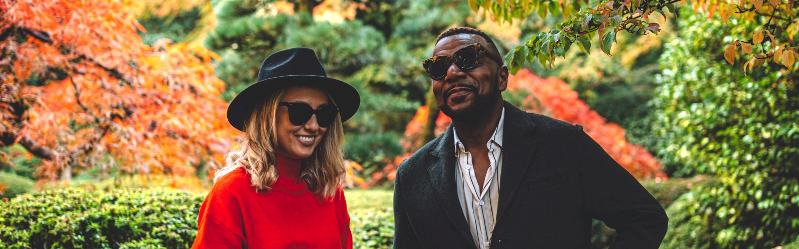 Two people are smiling, standing in a lush garden with vibrant autumn foliage, wearing stylish outfits and sunglasses.