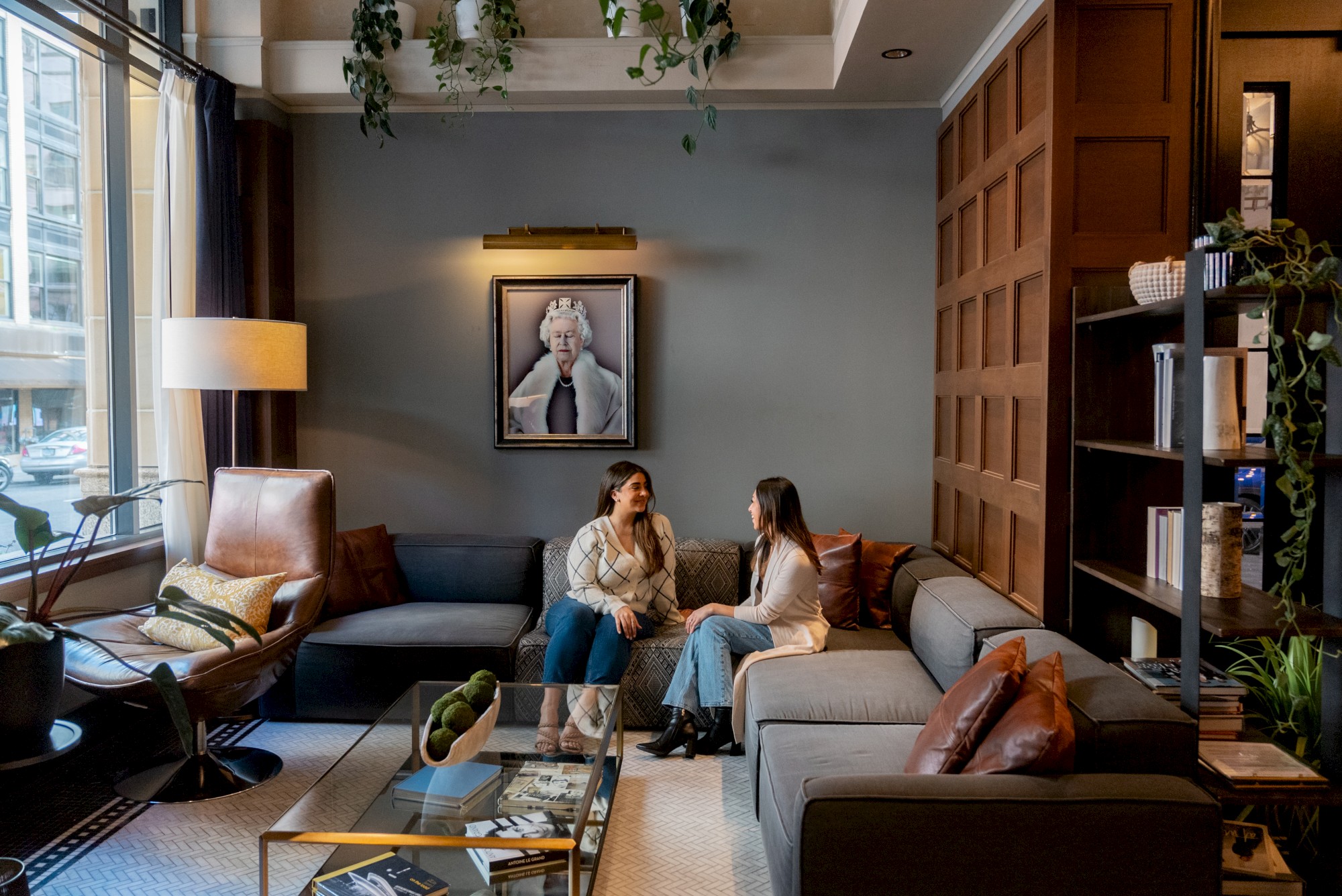 A cozy living room with two people sitting on a sectional sofa having a conversation, surrounded by books, plants, and a portrait on the wall.