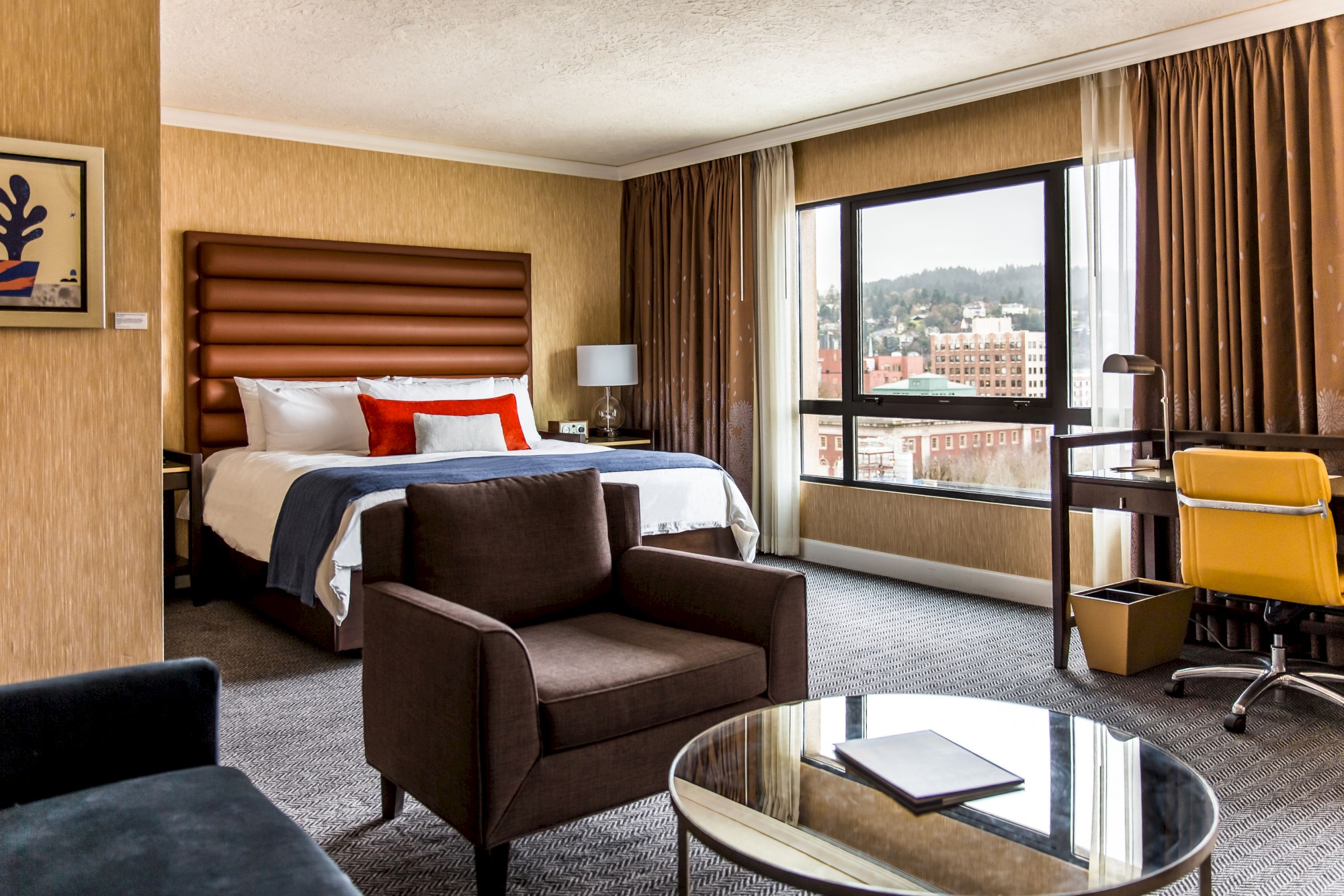 A modern hotel room with a bed, armchair, desk, and large window overlooking a cityscape, featuring warm tones and contemporary decor.