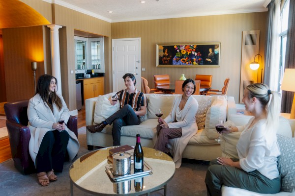 Four people are sitting on couches, enjoying wine in a cozy living room with warm lighting and decorative artwork on the wall.