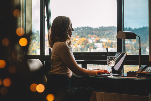 A person is sitting at a desk by a window, working on a laptop, with scenic views outside and a lamp nearby.