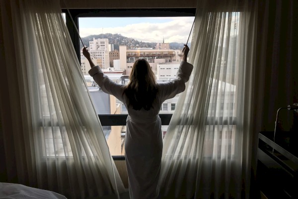 A person in a robe opens curtains, revealing a cityscape view with distant buildings and hills visible through a large window.