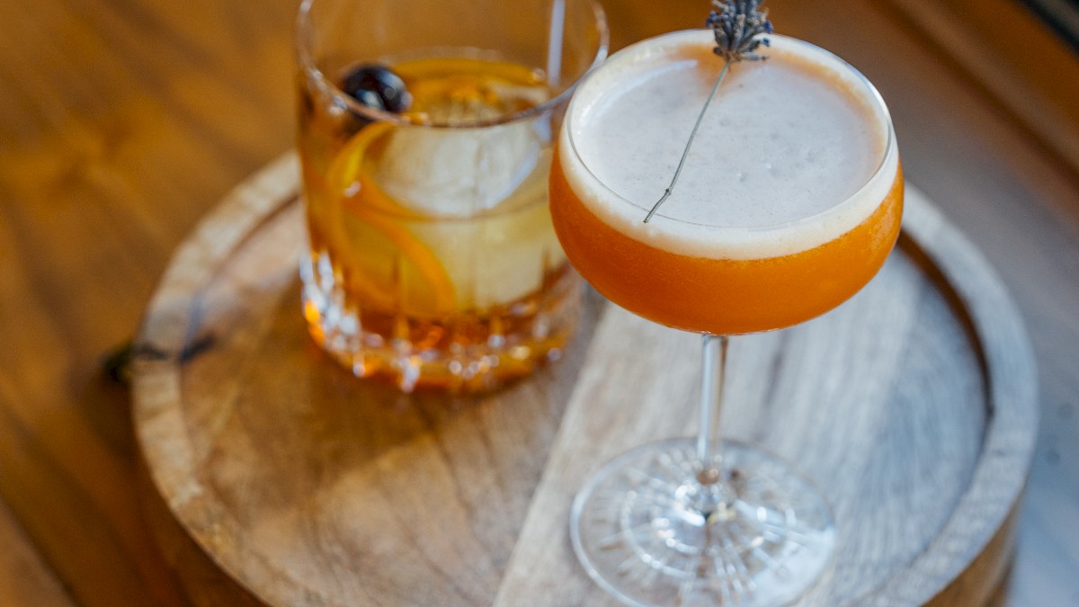 Two cocktails on a wooden tray: an old-fashioned with a cherry and an orange drink in a coupe glass garnished with lavender.