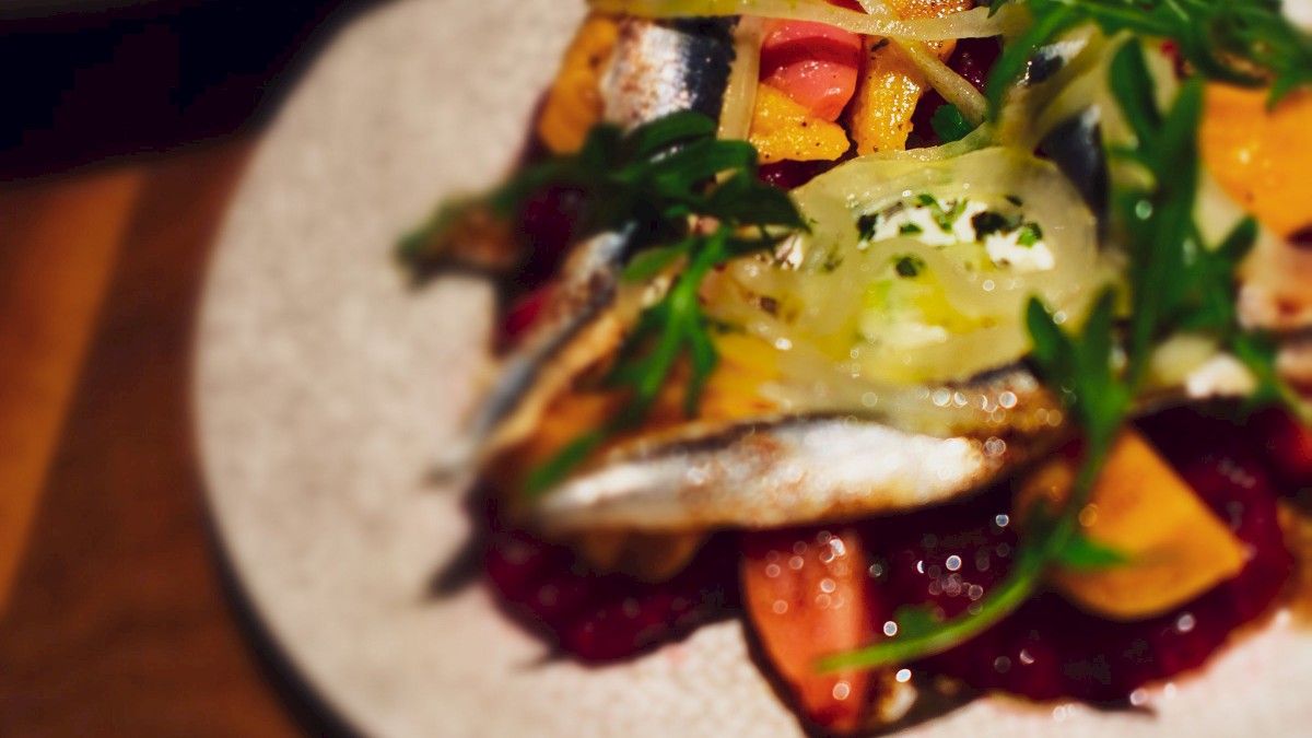 A dish of grilled fish with vegetables and arugula on a plate, with another dish blurred in the background.