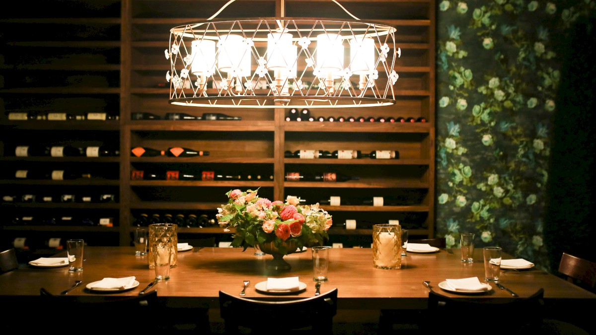 A dining table set with plates and glasses in a dimly lit room, with a floral centerpiece and a wall of wine bottles.