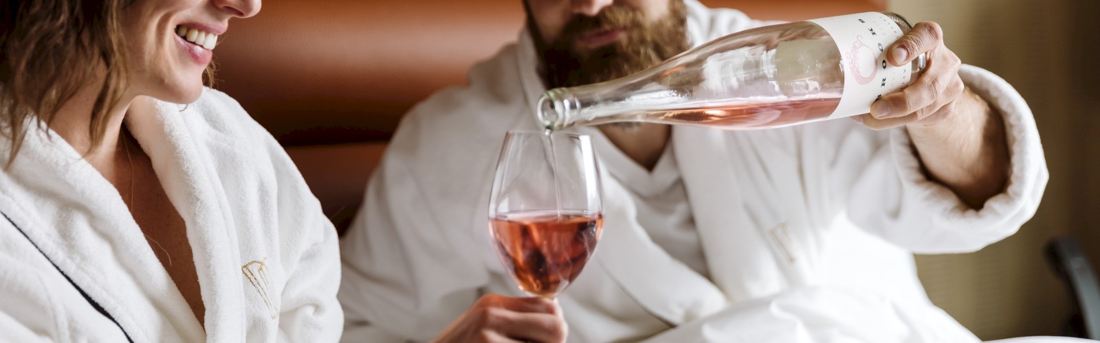 A couple in white bathrobes relax on a bed, with the man pouring rosé wine into a glass held by the woman, both smiling contentedly.