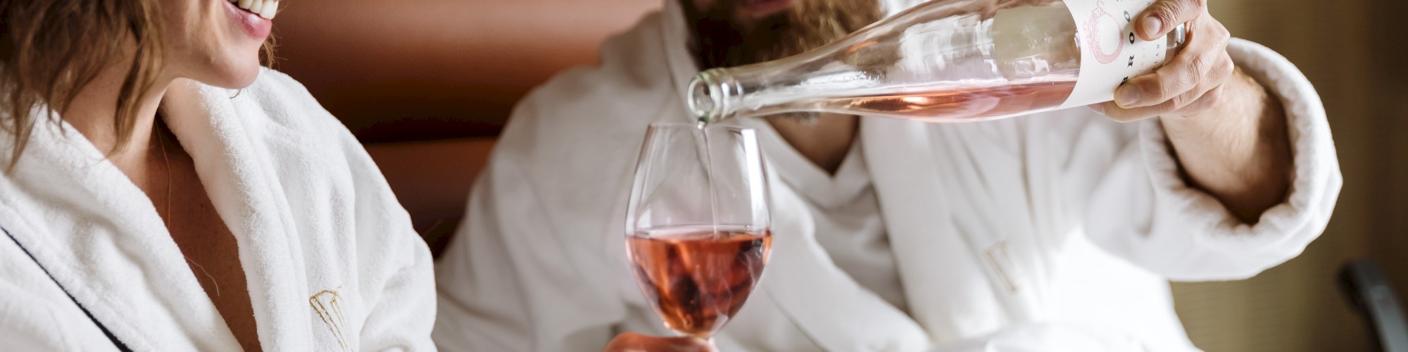 A couple in white bathrobes relax on a bed, with the man pouring rosé wine into a glass held by the woman, both smiling contentedly.