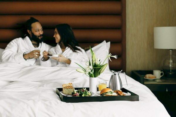 A couple in white robes enjoys breakfast in bed in a hotel room. A tray with food, flowers, and drinks is on the bed.