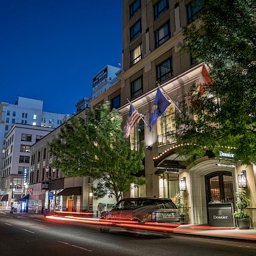 A city street at night with cars, a building with flags, and light trails from moving vehicles.