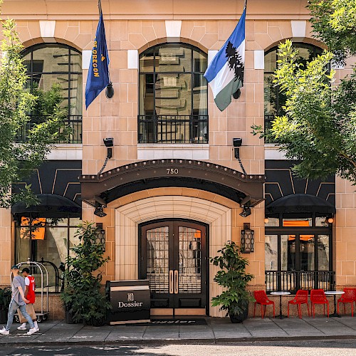 An elegant building entrance with tall arched windows, flags, outdoor seating, trees, and a person walking by on the sidewalk.