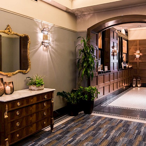 The image shows a stylish hallway with a wooden dresser, ornate mirror, decorative plants, and elegant lighting fixtures, leading to a corridor.