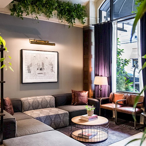 A cozy living room with a sectional sofa, plants, framed art, coffee tables, and natural light from large windows with dark curtains.