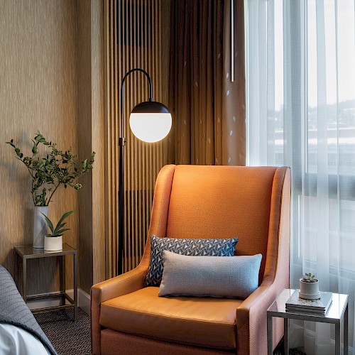 A cozy room corner featuring an orange armchair with pillows, a side table, a lamp, and a window with sheer curtains.