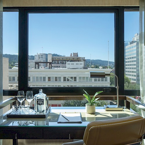 A hotel room desk with a view of city buildings, featuring glasses, a water bottle, a plant, and a lamp.