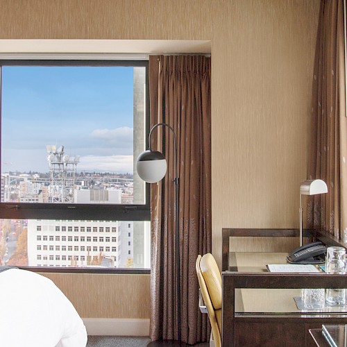 A hotel room with large windows showing a cityscape, containing a bed, desk, chair, lamp, and phone, creating a cozy atmosphere.