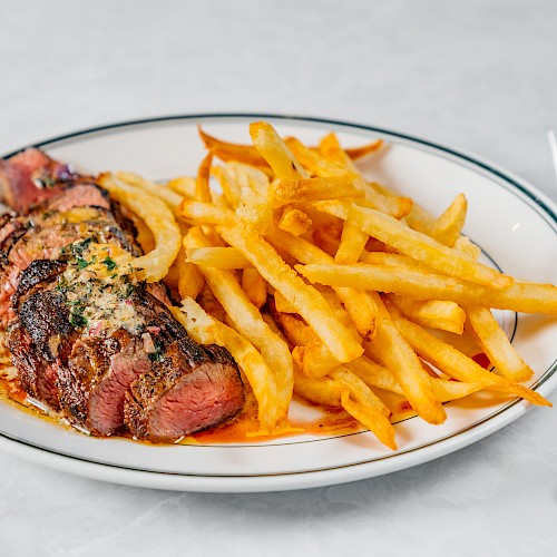 A plate with sliced steak topped with herbs and butter, served alongside crispy French fries, with a fork and knife beside it.