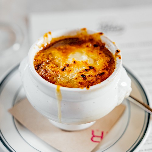 A bowl of French onion soup with melted cheese, placed on a saucer with a napkin and spoon, atop a menu or paper.