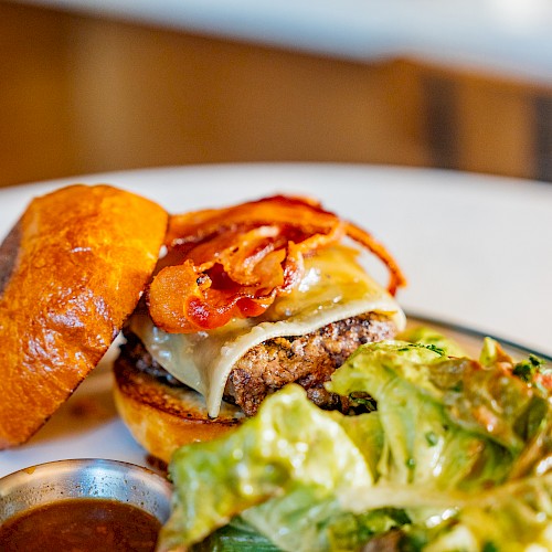 A cheeseburger with bacon on a plate, served with a side salad and a small dish of sauce.